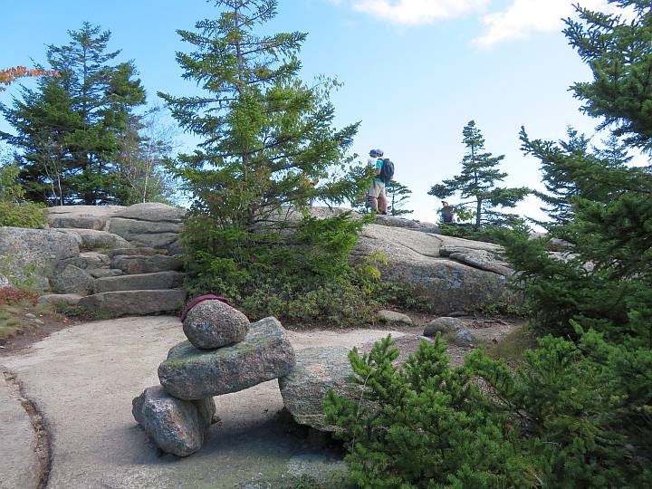 Large cairn trail marker and two hikers standing on a large rock next to trees.