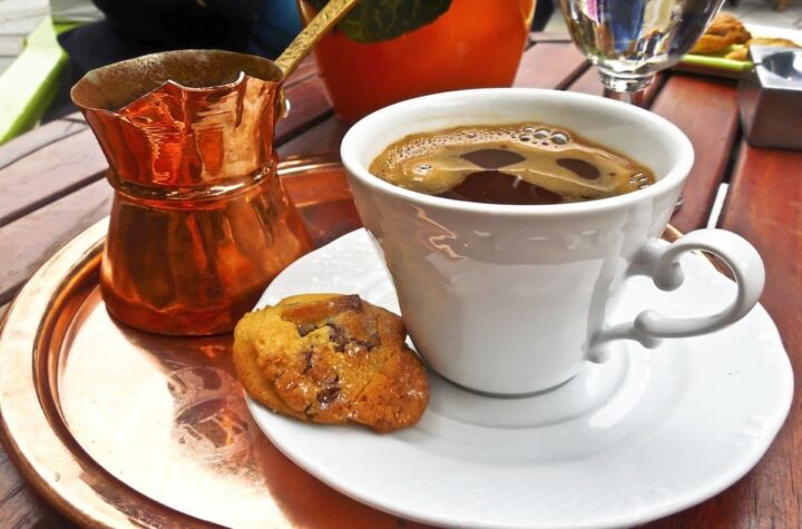 Traditional Greek coffee in a white cup and saucer with a cookie on the saucer next to a briki.