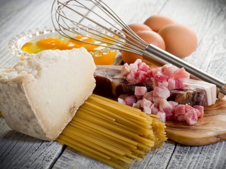 Raw ingredients for carbonara: eggs, pork jowls, a block of Pecorino Romano cheese, plus dry spaghetti pasta, and a whisk.