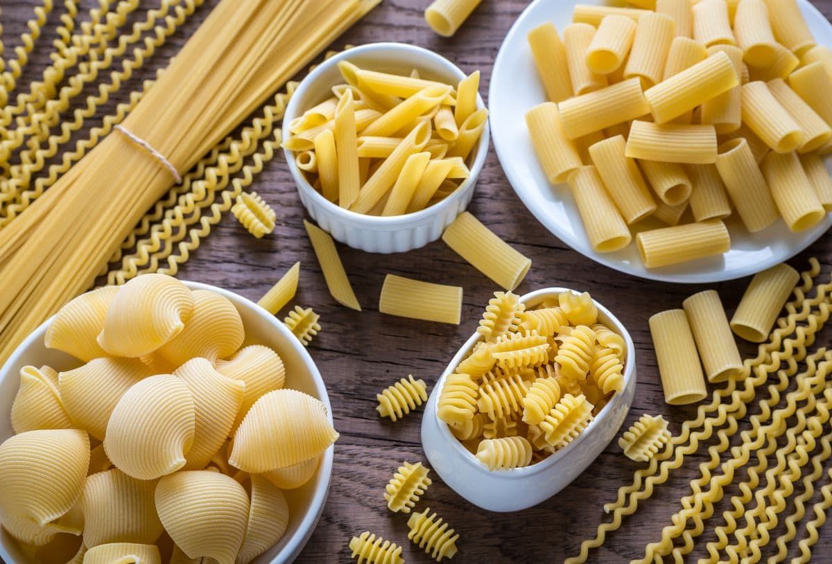 An assortment of uncooked dry pasta shapes displayed in small bowls on a wooden table.