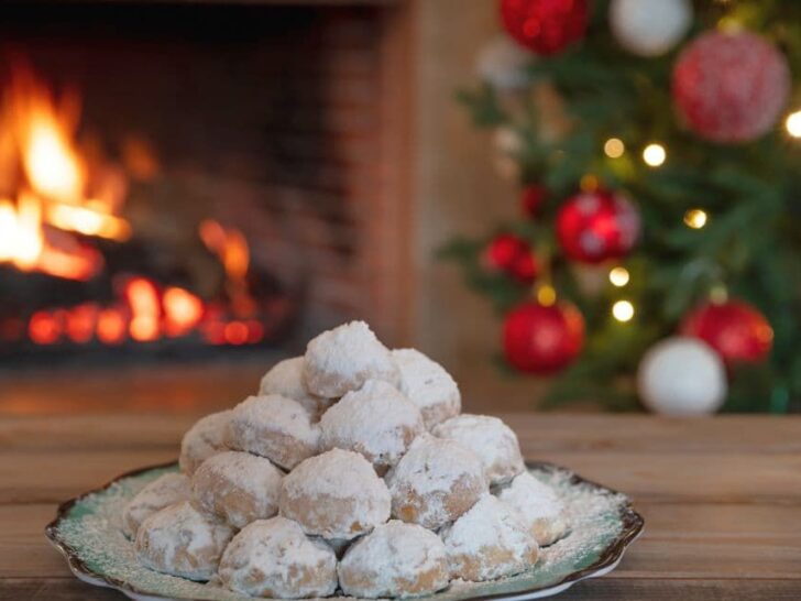 Greek Christmas cookies kourabiedes on a plate with fireplace and Christmas tree blurred in background.