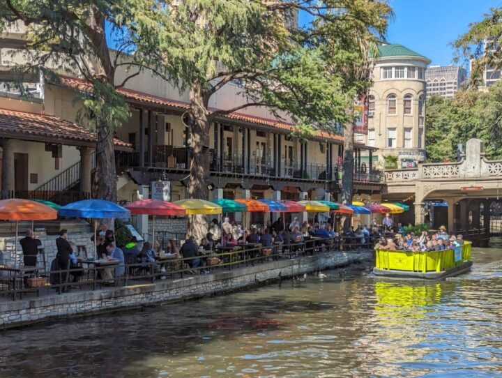 San Antonio River Walk with river taxi and people dining al fresco under colorful umbrellas.