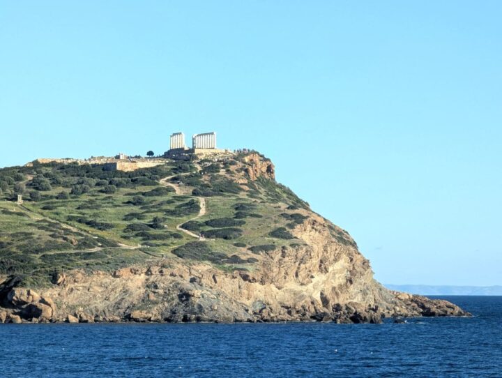 Long distance view of Temple of Poseidon atop a cliff overlooking the Aegean Sea on a sunny day with calm sea.