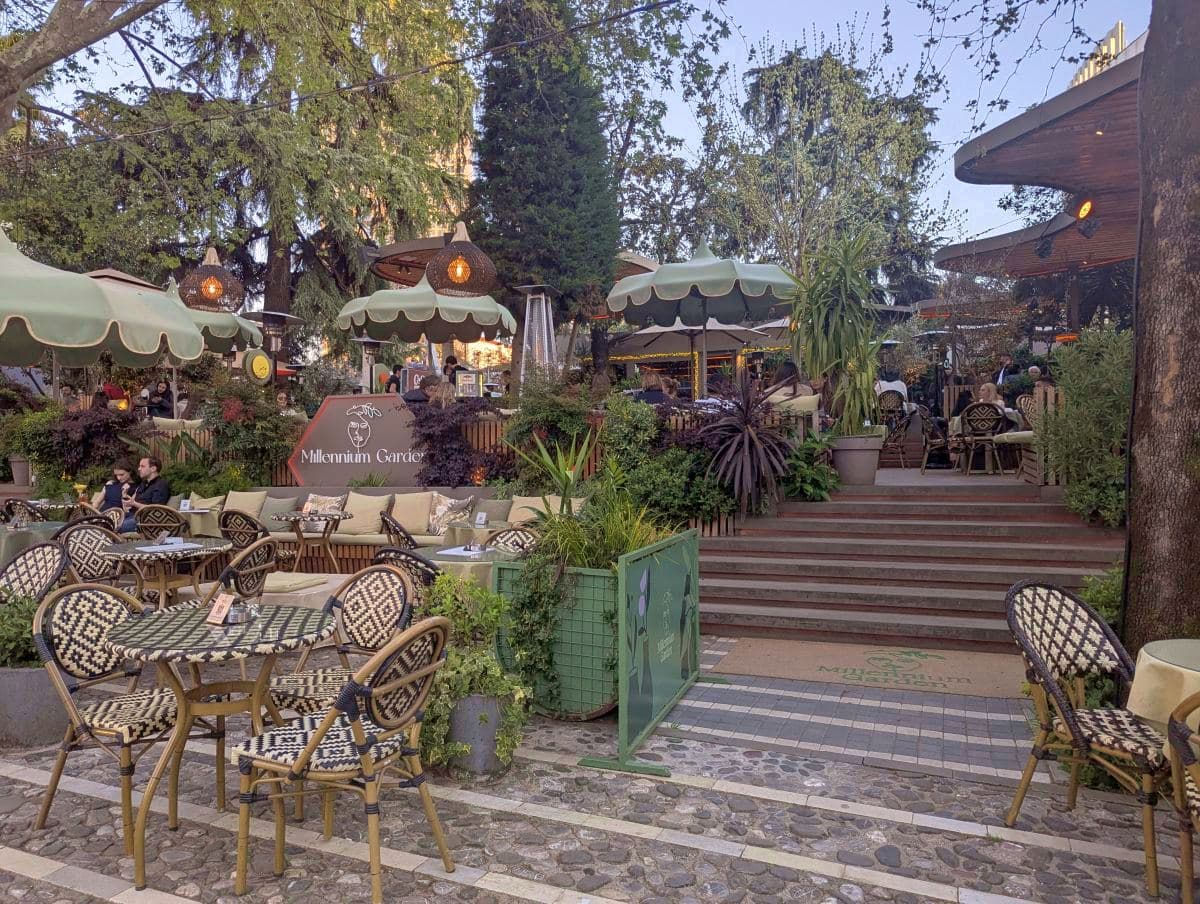 The outdoor patio of the Millennium Garden café in Tirana, featuring patterned bistro chairs, cozy lounge seating, and scalloped umbrellas nestled among lush trees and green plants.