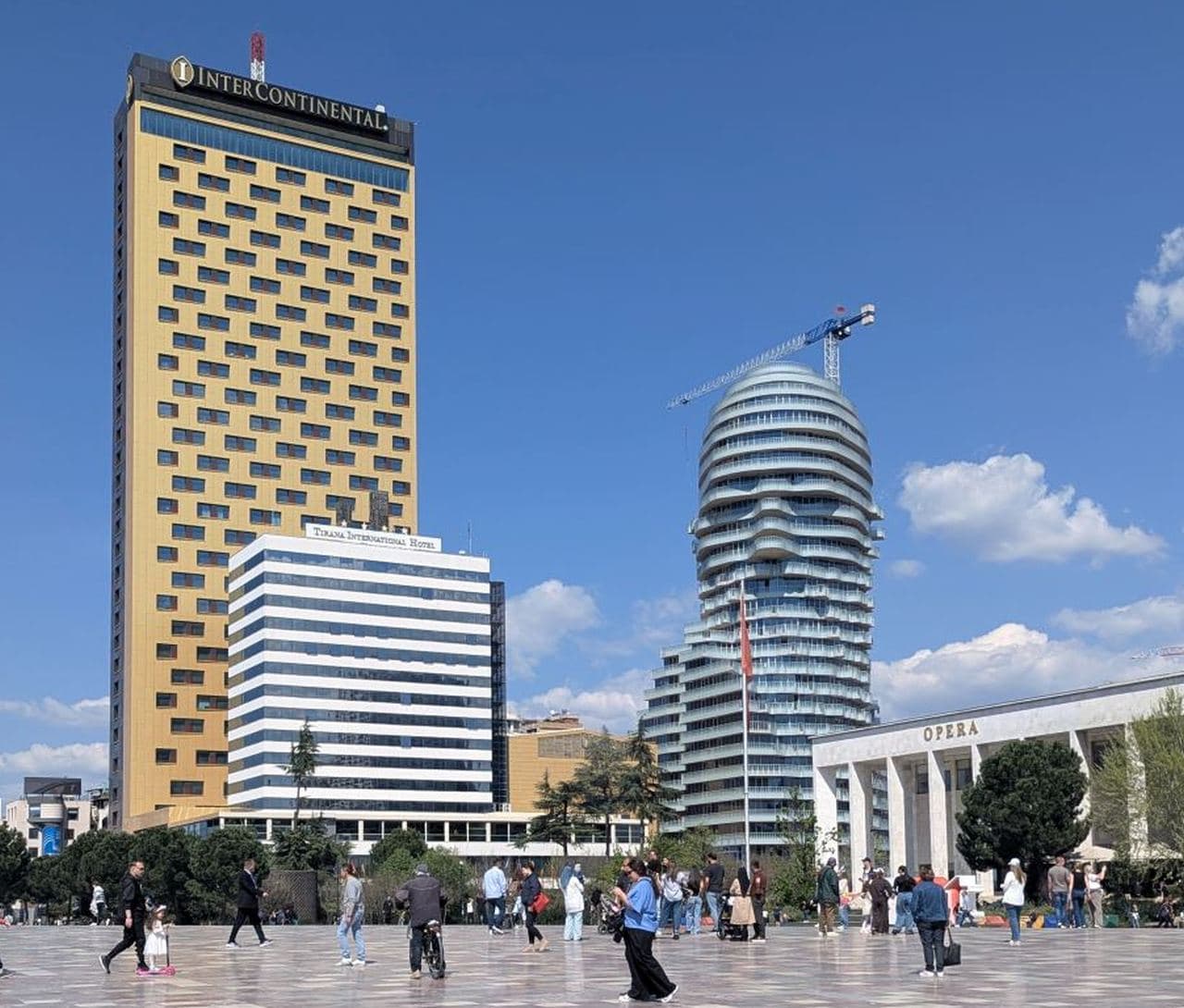 Pedestrians cross the paved Skanderbeg Square in Tirana, Albania, with the face-shaped Tirana’s Rock skyscraper and golden InterContinental Hotel under a blue sky.