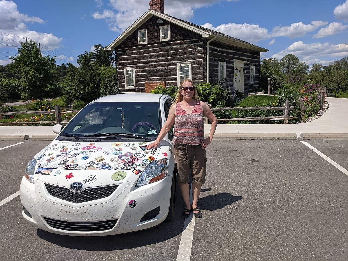 A woman standing next to white Toyota Yaris sedan with dozens of stickers on the hood, in front of a log cabin in Mississauga Ontario, Canada.