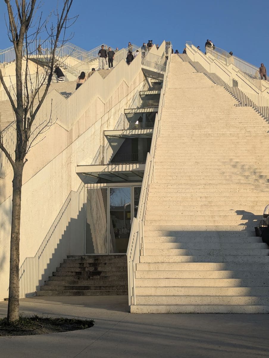 Visitors climb the steep, modern concrete steps of the renovated Pyramid of Tirana under a clear blue sky during a sunny day in Albania.